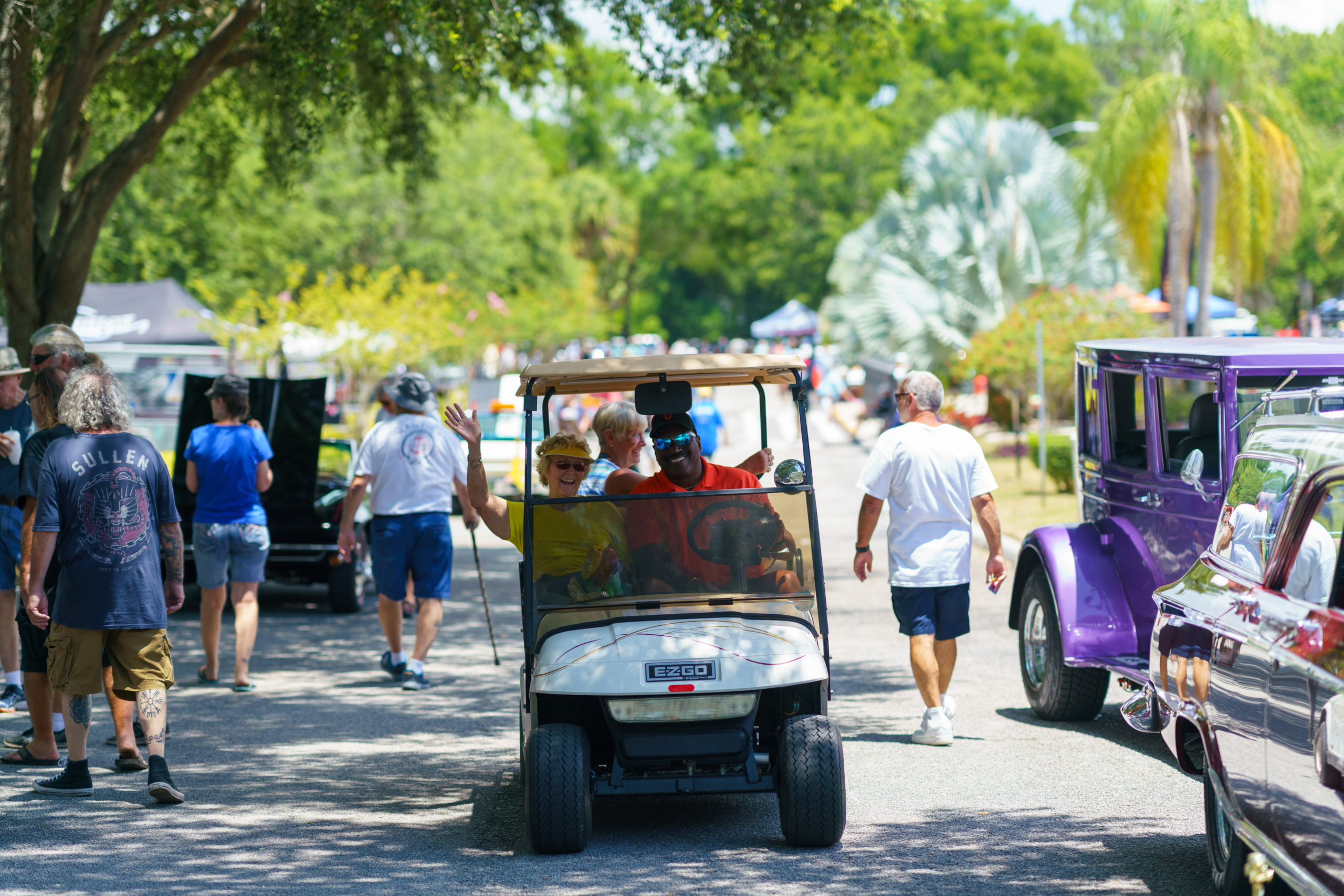 A crowd of people walking down a road lined with classic cars and hot rods, in the middle a golf cart sitting two people who are smiling and joyful as they ride past.