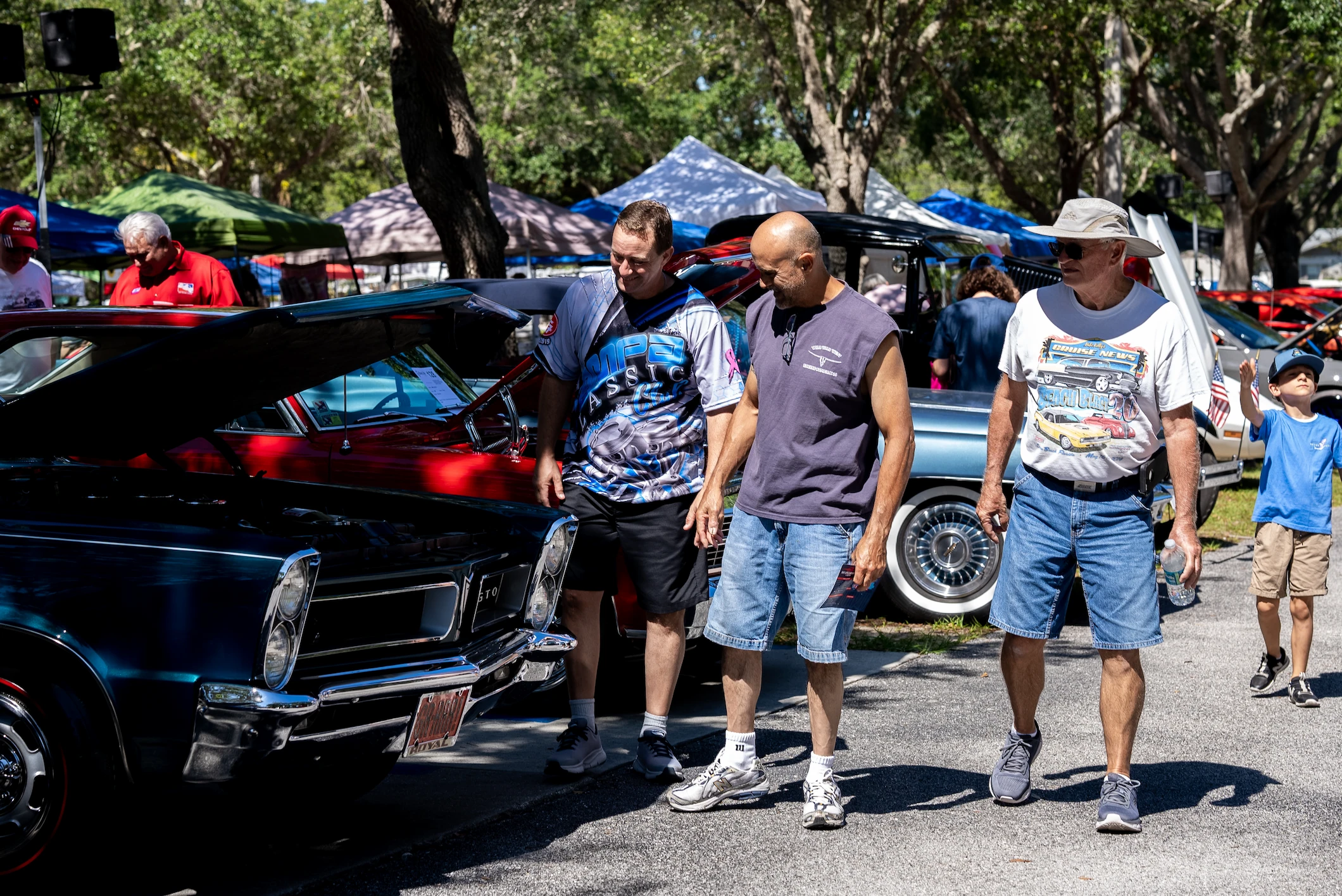 A crowd of people walking down a road lined with classic cars and hot rods, in the middle a golf cart sitting two people who are smiling and joyful as they ride past.
